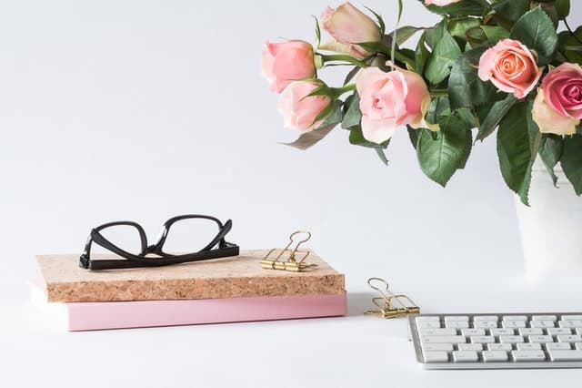 A chic and minimalist work desk setup with a bouquet of pink roses, stylish eyeglasses resting on top of a cork notebook, elegant gold paper clips, and a sleek white keyboard.