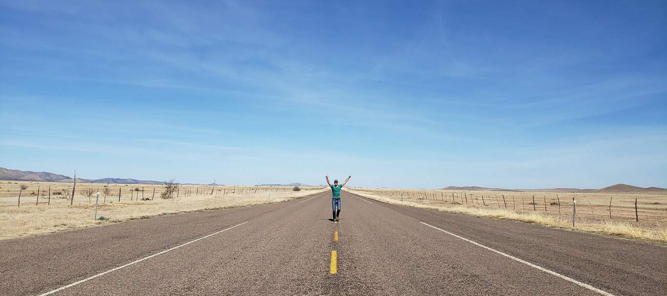 A person stands in the middle of a long, empty road with arms joyfully stretched towards the expansive blue sky, surrounded by the tranquility of a flat, open landscape.