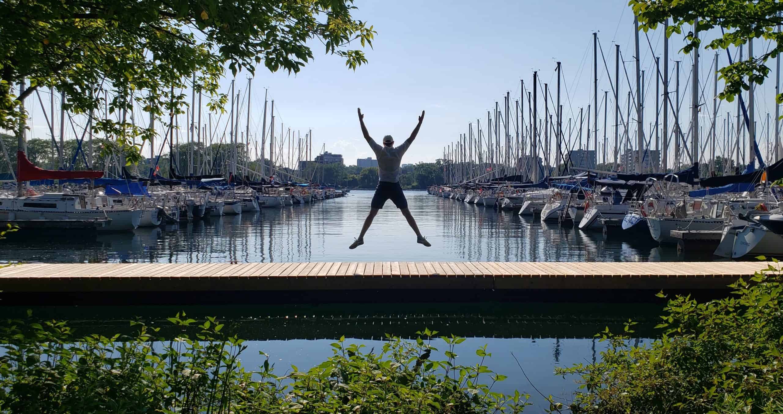 Person celebrating joyfully with an exuberant jump in front of a serene marina lined with sailboats on a beautiful sunny day.
