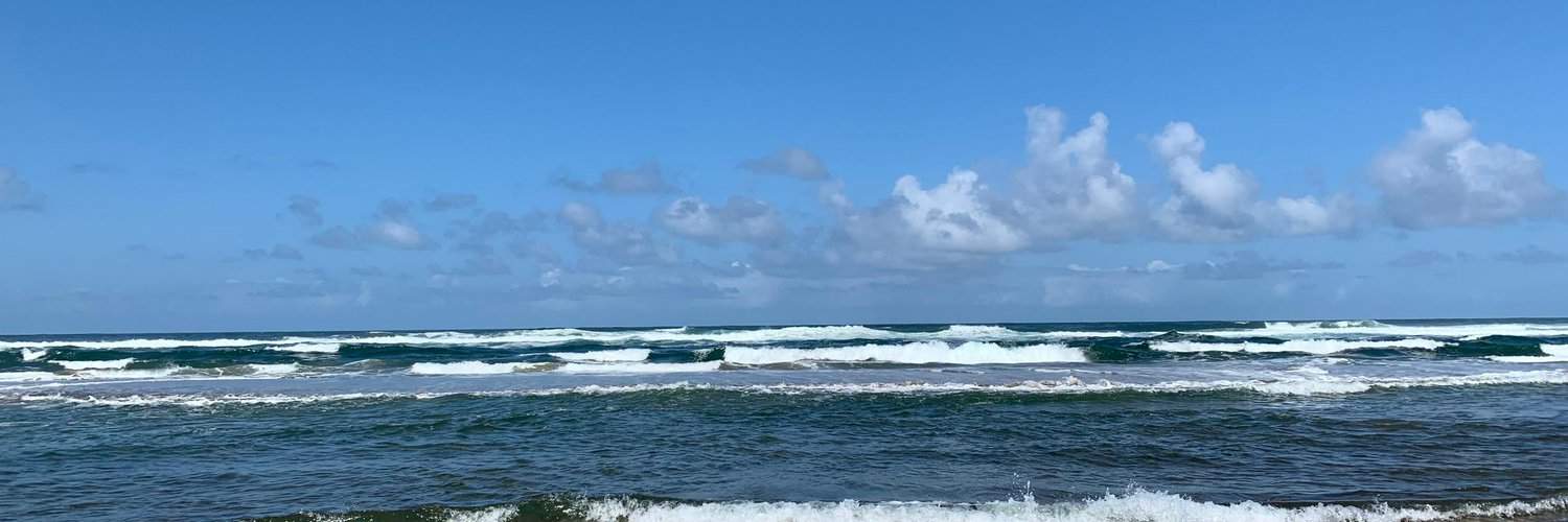 Expansive blue ocean under a wide sky with scattered clouds, waves rolling towards the shore in a serene rhythm.