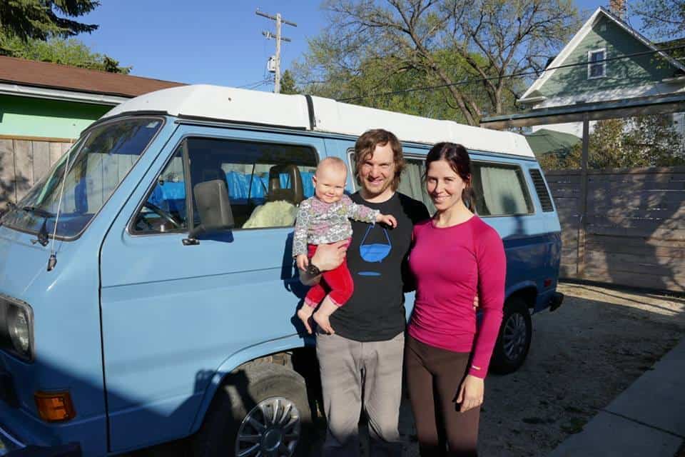 A happy family with a baby smiling near their blue camper van, ready for an adventurous road trip.
