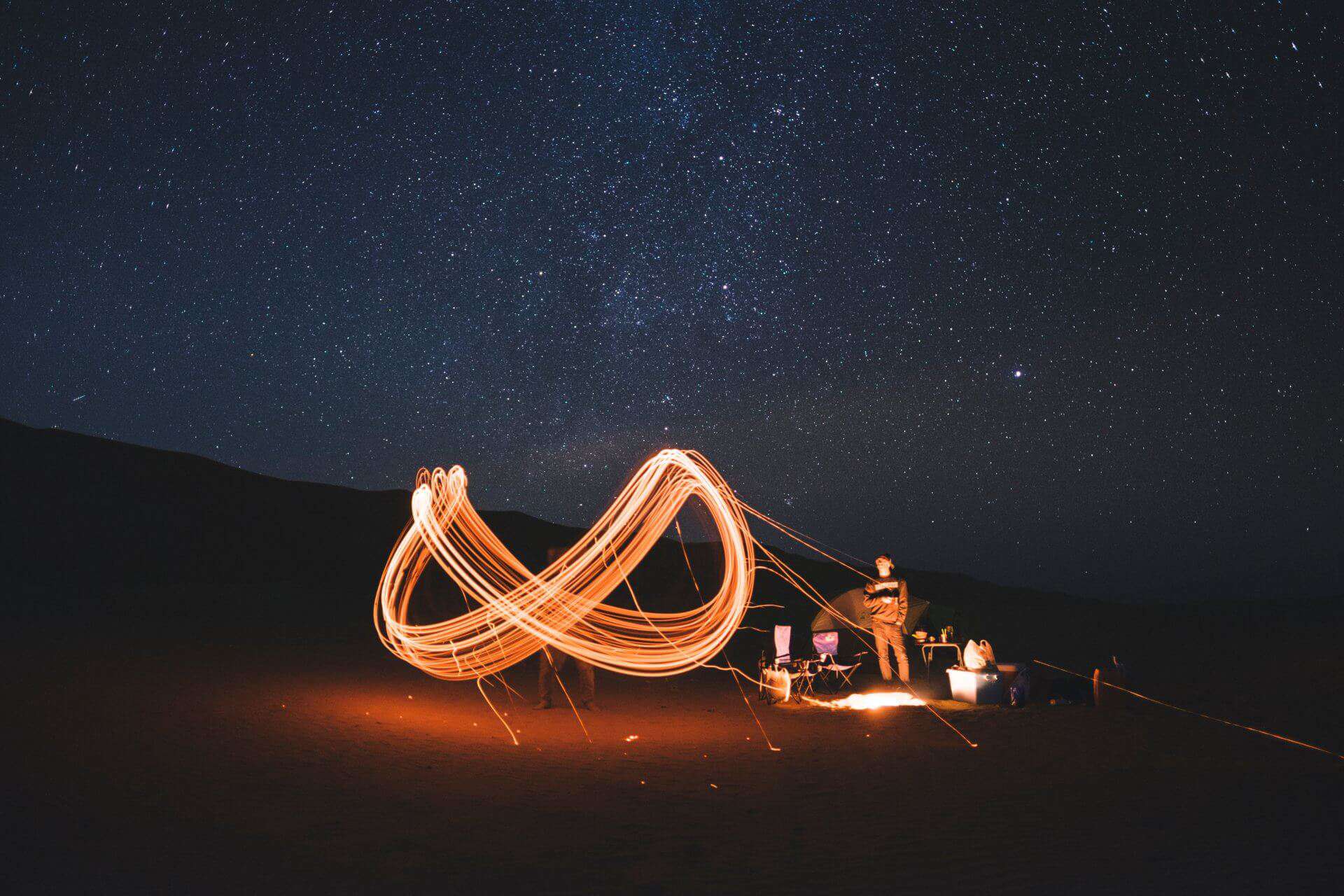 A lone camper under a starry sky creates a light trail with a spinning device, lending a magical atmosphere to a peaceful night in the wilderness.