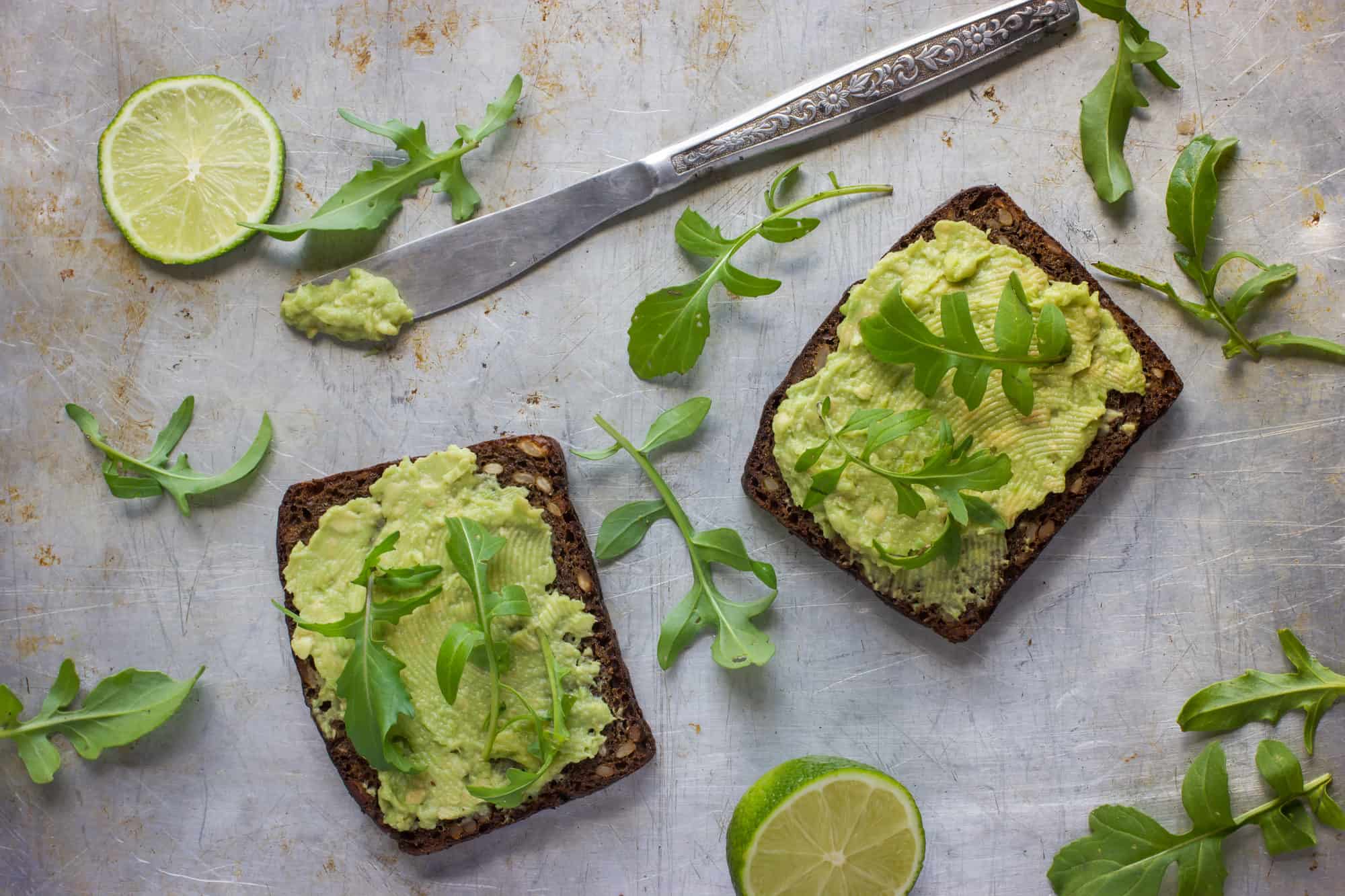 Two slices of avocado toast garnished with lime and fresh herbs on a rustic metal surface.