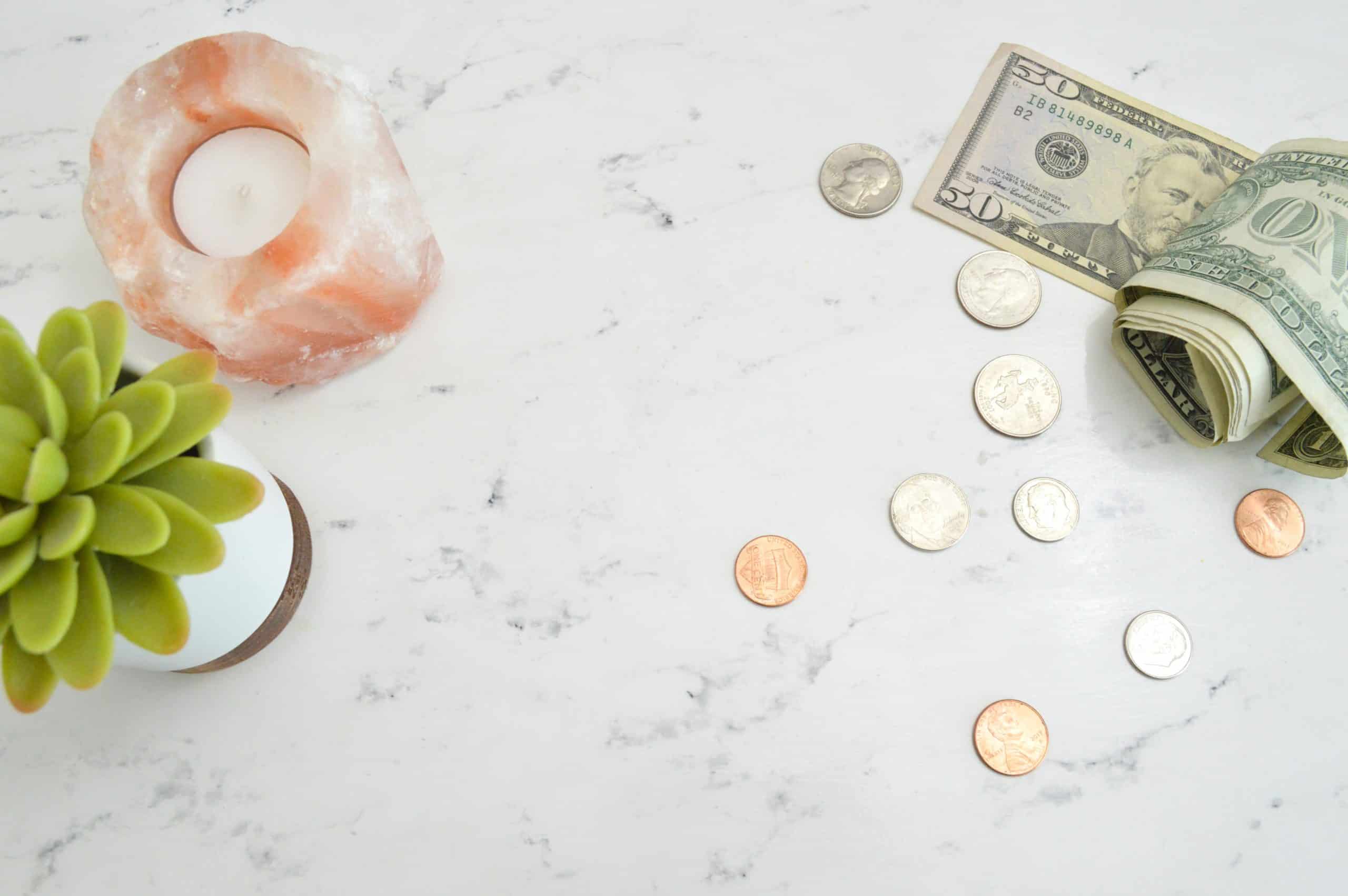 A serene marble countertop with a soothing arrangement of a pink salt candle holder, a potted succulent, and scattered us currency including coins and rolled bills.