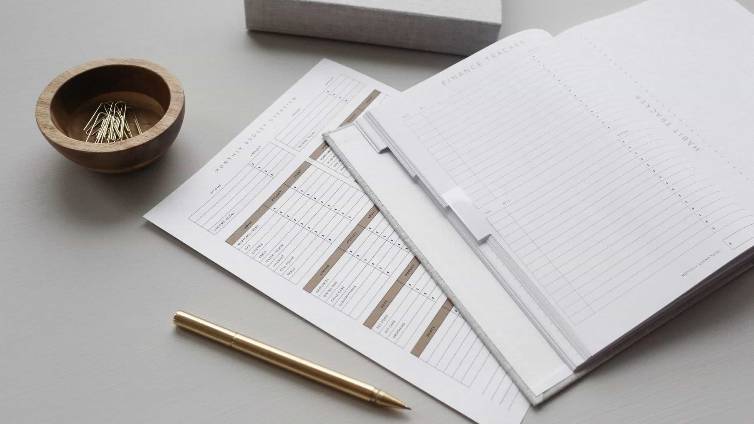 A neatly organized workspace featuring an open planner, a collection of financial documents, a wooden bowl with paper clips, and a gold pen, all resting on a gray desk surface.