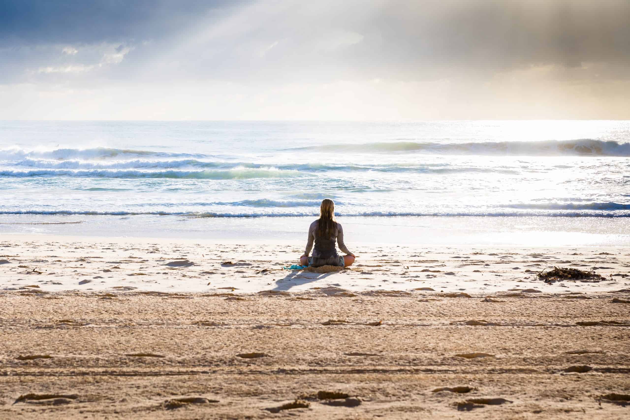 A tranquil moment as a person sits on a sandy beach, gazing out at the serene waves under a soft golden sunlight piercing through the clouds.