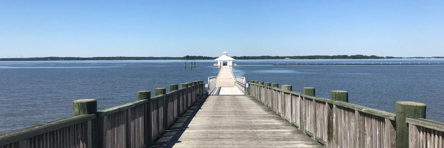 A serene wooden pier leading out into a calm blue expanse of water, crowned by a small white gazebo under a clear sky.