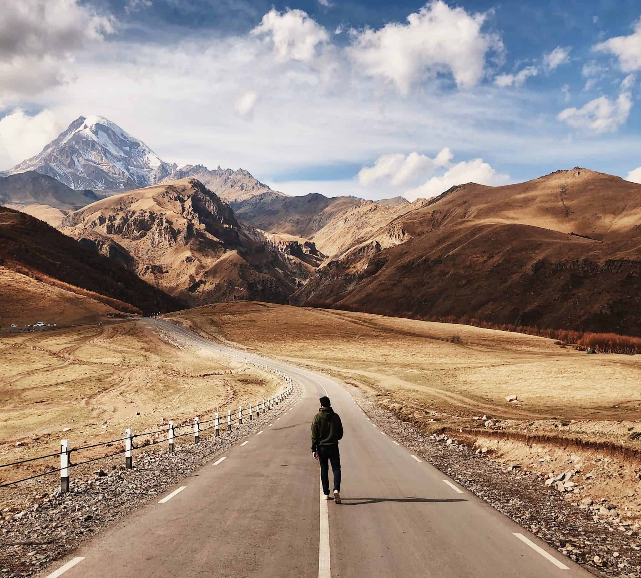 A lone traveler walks down an open road stretching through a majestic mountainous landscape under a vast open sky.