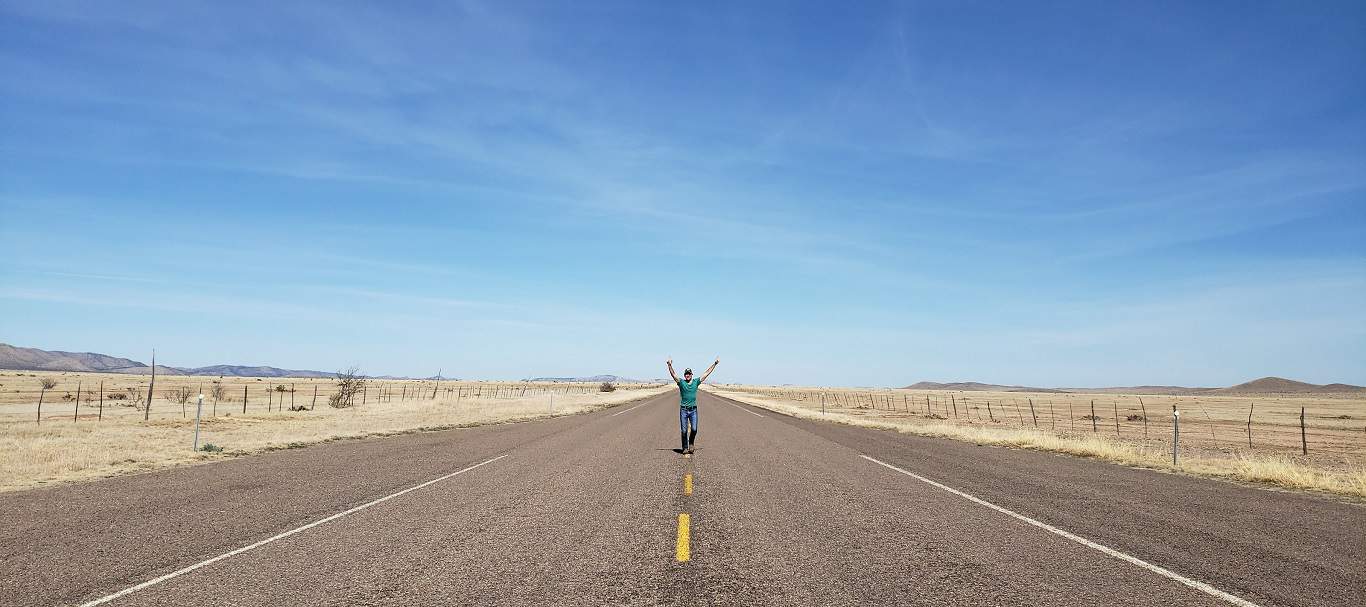 A person celebrating freedom with arms raised in the middle of a vast, empty road stretching toward the horizon under a clear blue sky.