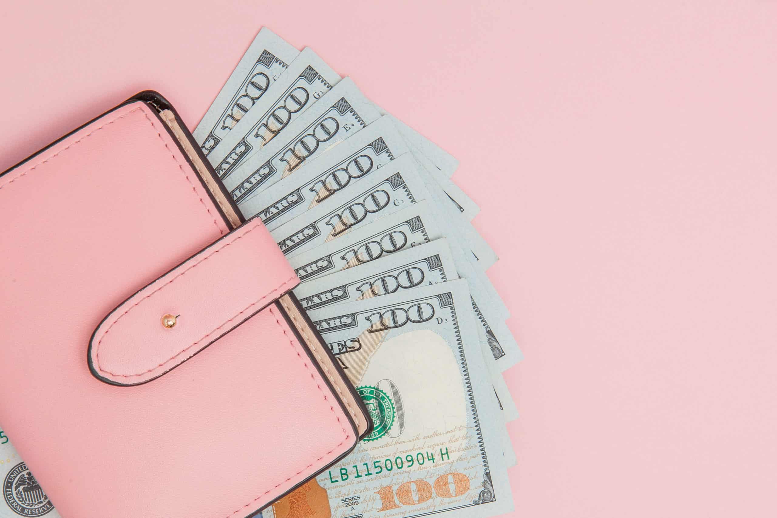 A pink wallet overflowing with multiple one hundred us dollar bills on a soft pink background.