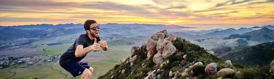 A joyful man giving a thumbs up while posing on a scenic mountaintop at sunset.