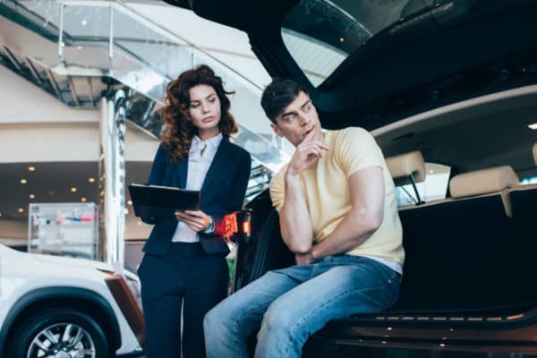 A sales consultant with a digital tablet discussing vehicle features with a thoughtful potential buyer sitting in the trunk of a new car at a dealership showroom.