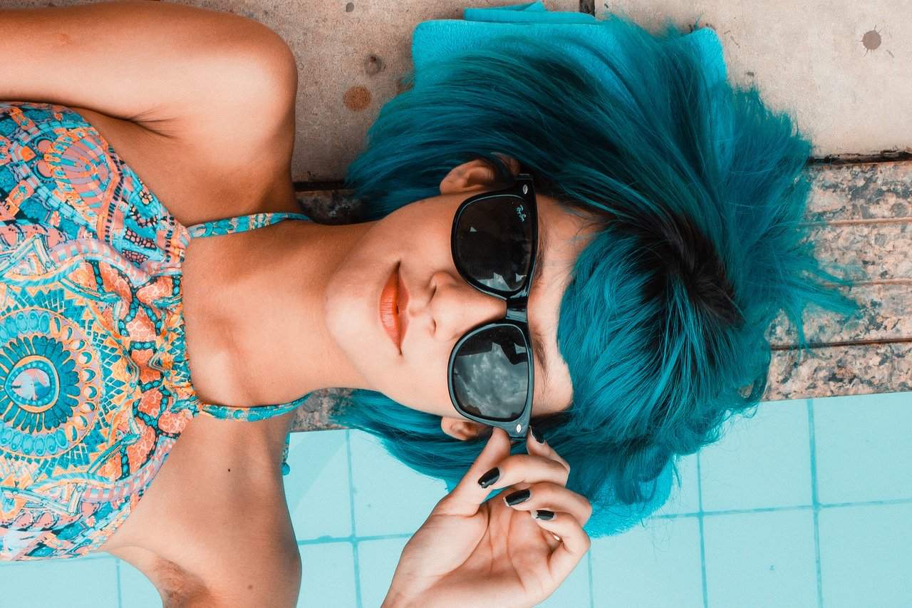A woman with vibrant blue hair relaxing poolside, peering over her sunglasses.