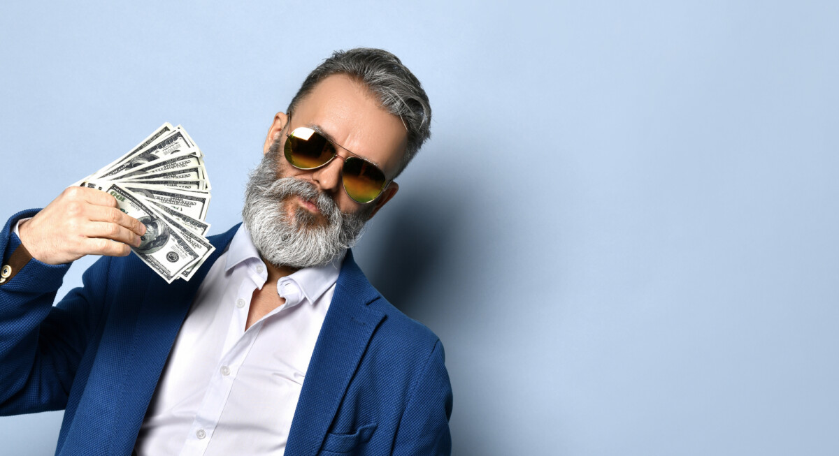 Gray-haired, bearded male in white shirt, jacket and sunglasses. He is showing a fan of hundred dollar bills in his hand, posing against blue studio background.