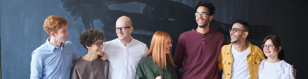 A diverse group of seven smiling individuals standing in front of a dark chalkboard, looking happy and confident, possibly coworkers or friends.