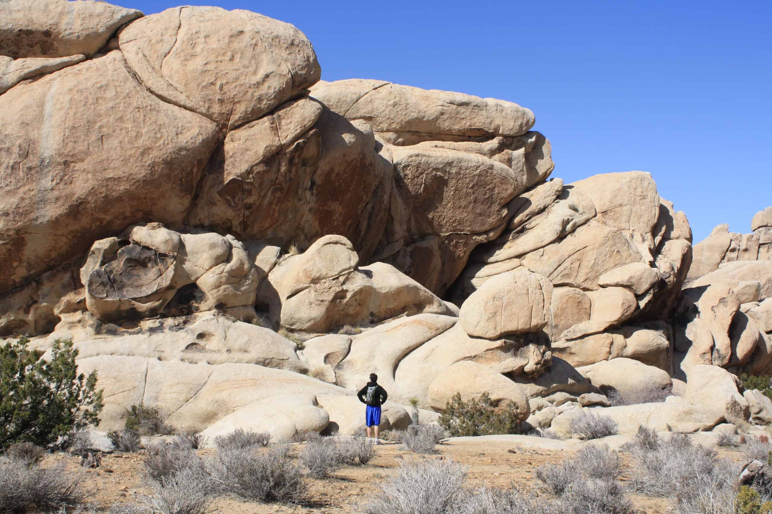 A lone explorer stands before a majestic landscape of towering boulders and rock formations under a clear blue sky.