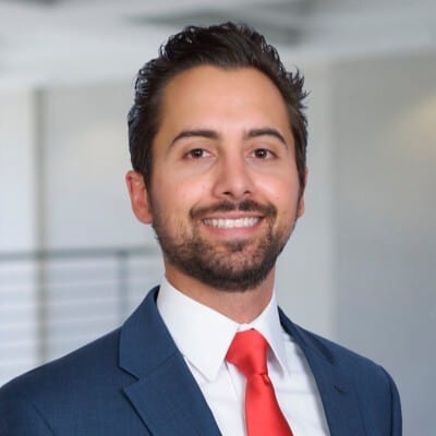 A professional man with a beard smiling confidently in a business suit with a red tie against an office background.