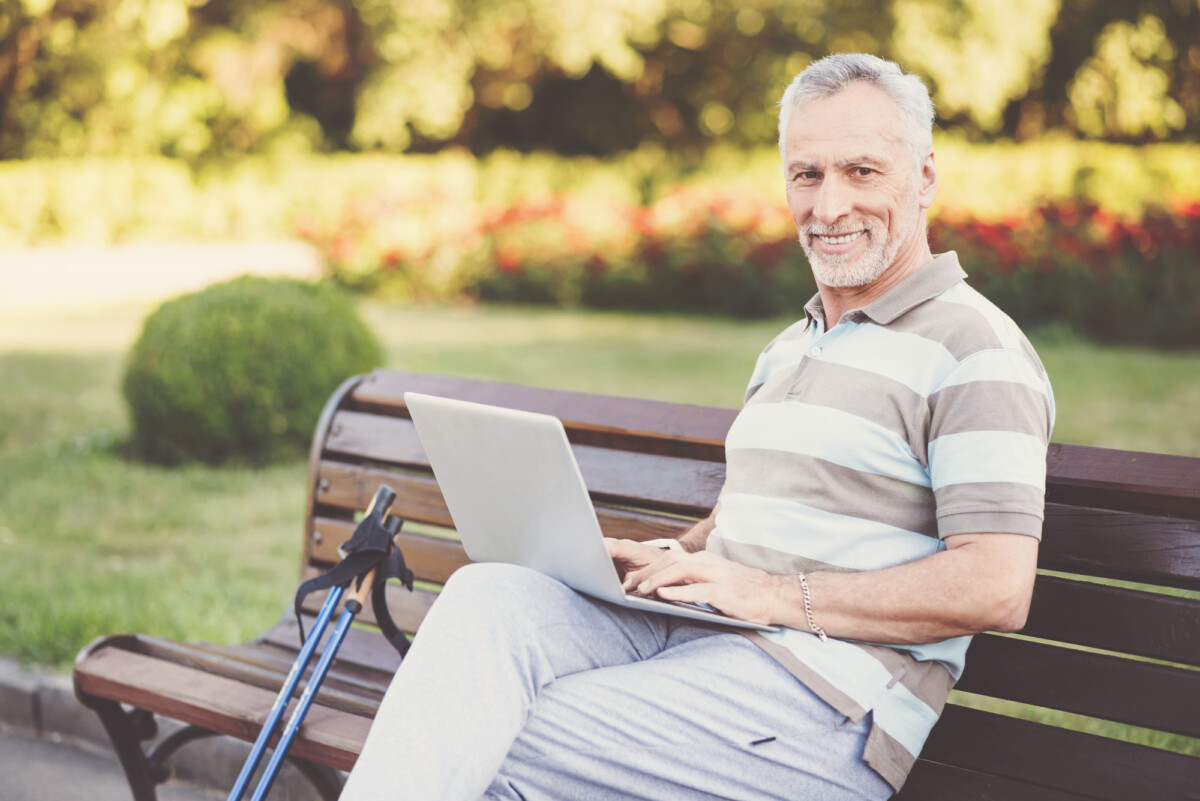 Cheerful good looking retiree sitting on the bench and using a laptop while working on it, enjoying working after retirement