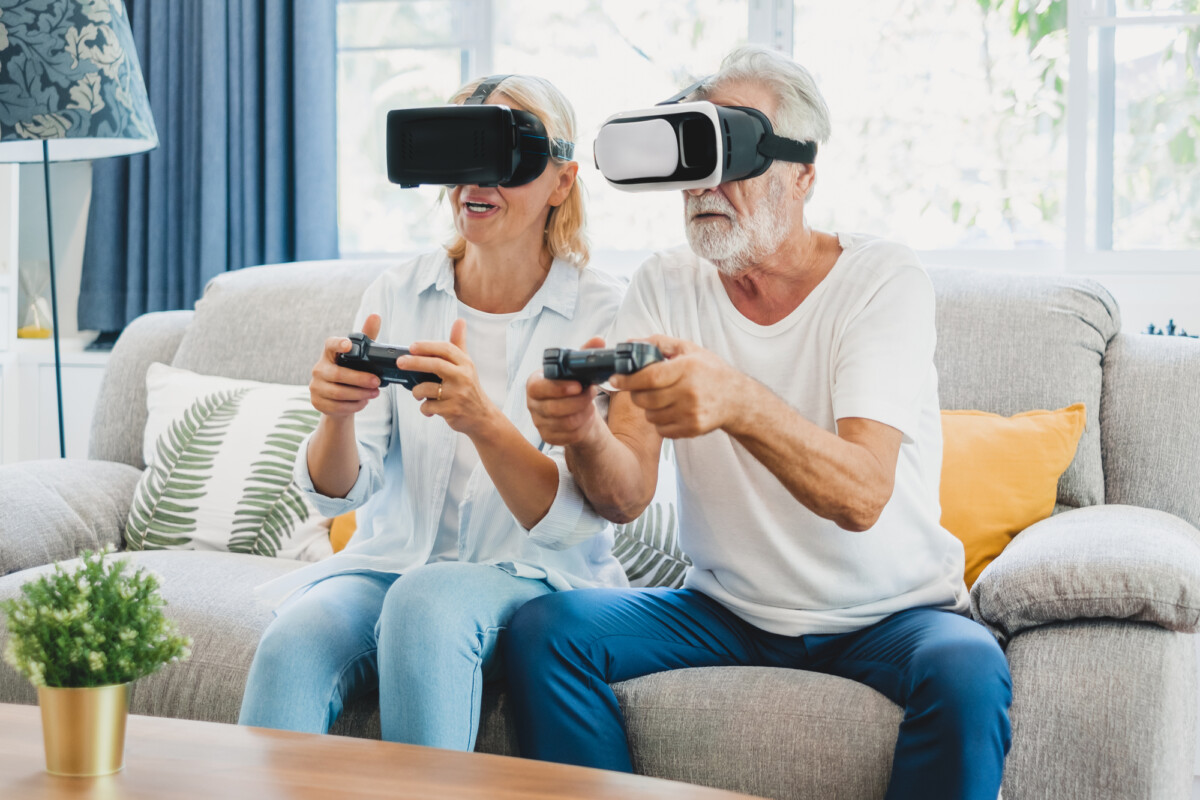 A retired husband and wife enjoy playing VR 3D games at home on couch.