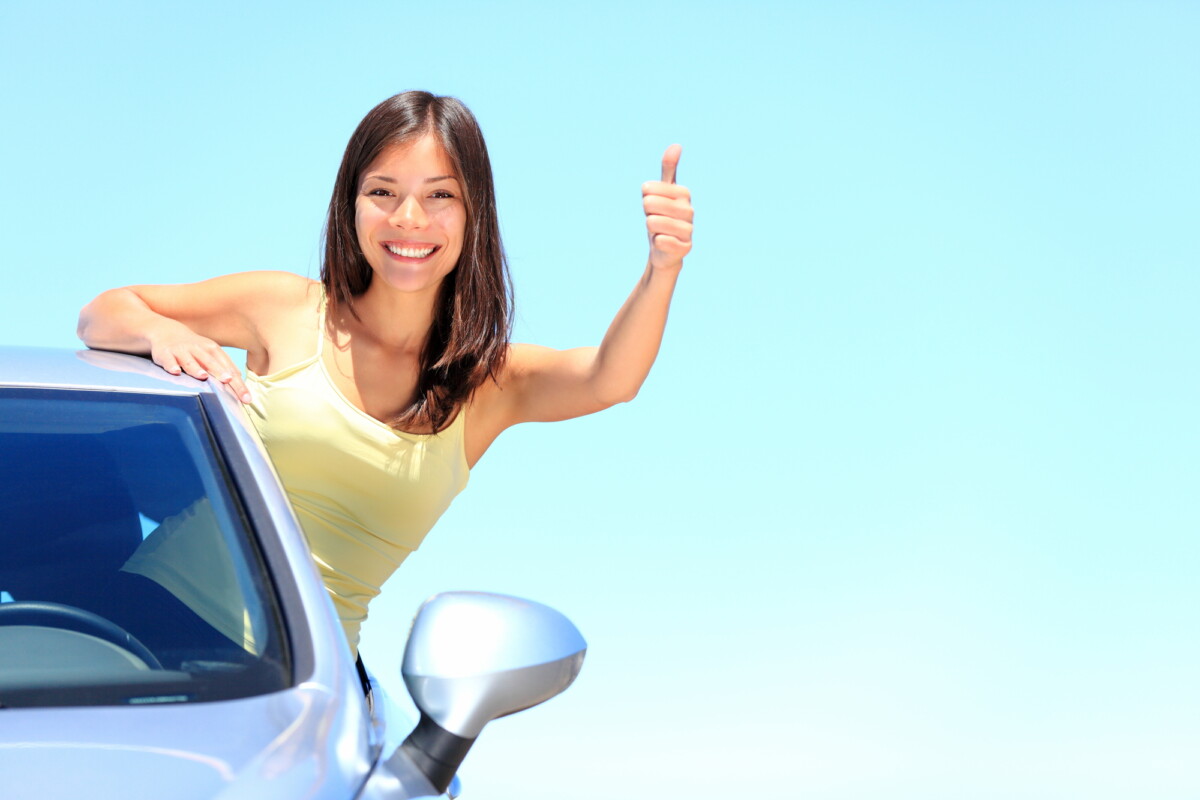 Car. Woman driver happy smiling showing thumbs up coming out of car window on blue summer sky above the clouds. Beautiful young mixed race Caucasian / Chinese Asian woman.