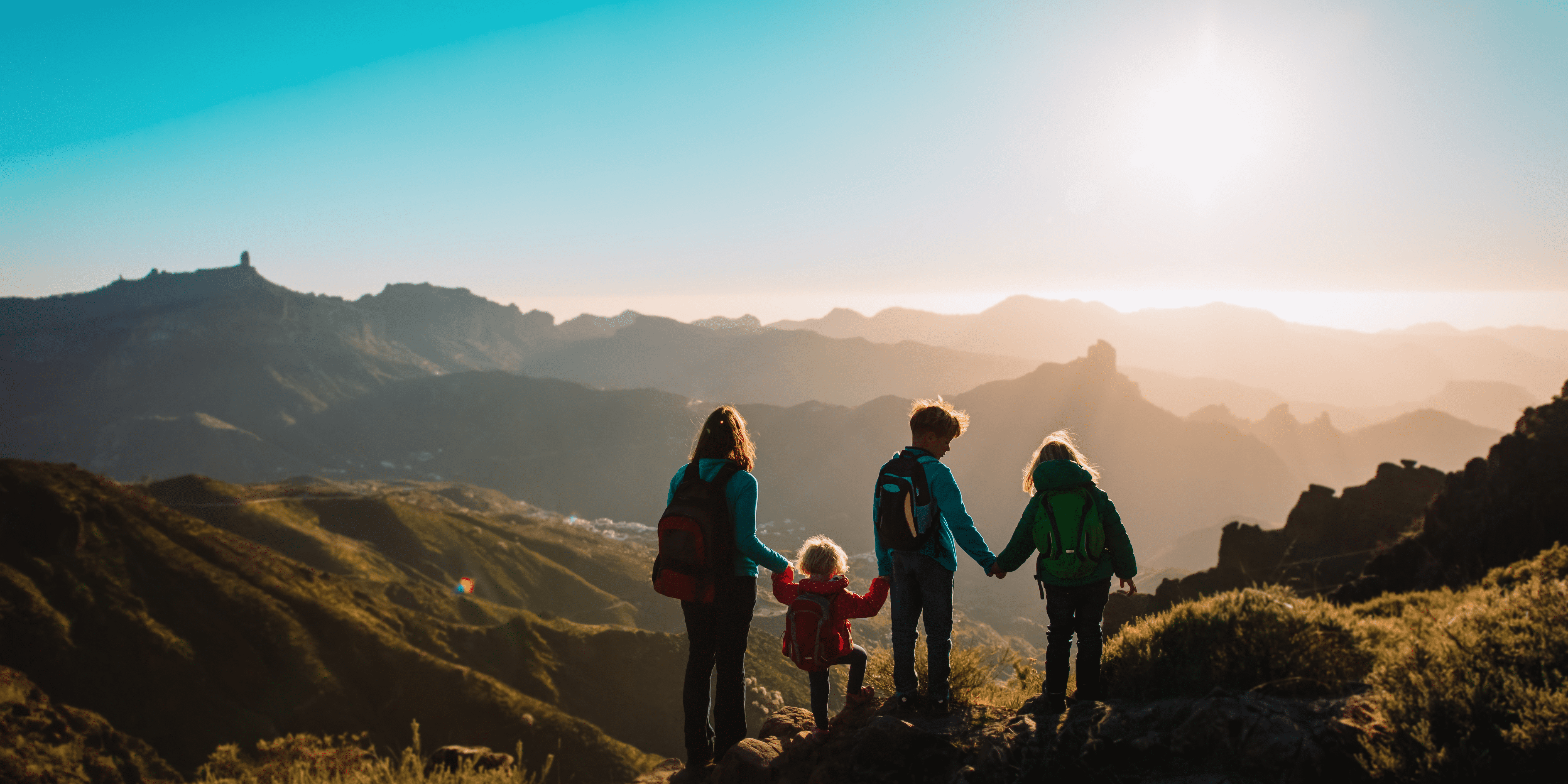 A family holding hands while standing on a mountain ridge, basking in the golden light of the setting sun with panoramic views of rugged peaks in the distance.