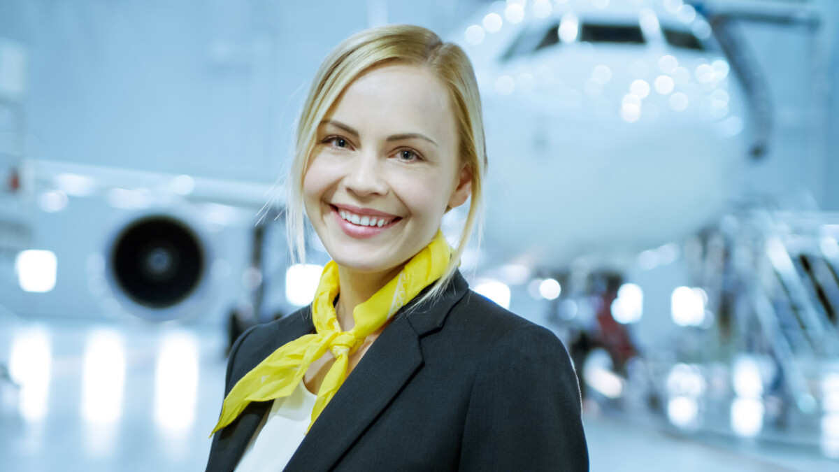 In a Aircraft Maintenance Hangar Young Beautiful Blonde Stewardess/ Flight Attendant Smiles Charmingly on Camera. In the Background Brand New Airplane is Visible.