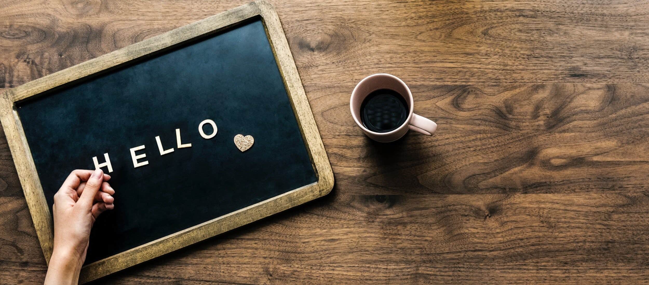 A person's hand arranging letter tiles spelling 'hello' on a chalkboard next to a heart symbol, with a cup of coffee on a wooden table, conveying a warm and welcoming message.