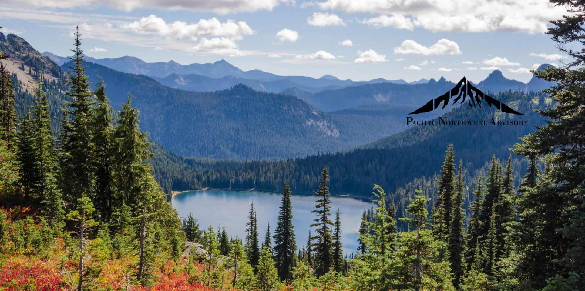 A serene mountain landscape with a clear blue lake nestled among forested hills under a partly cloudy sky.