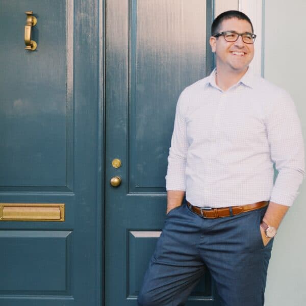 A man in a button-up shirt and glasses standing with a confident smile beside a dark blue door with brass fixtures.