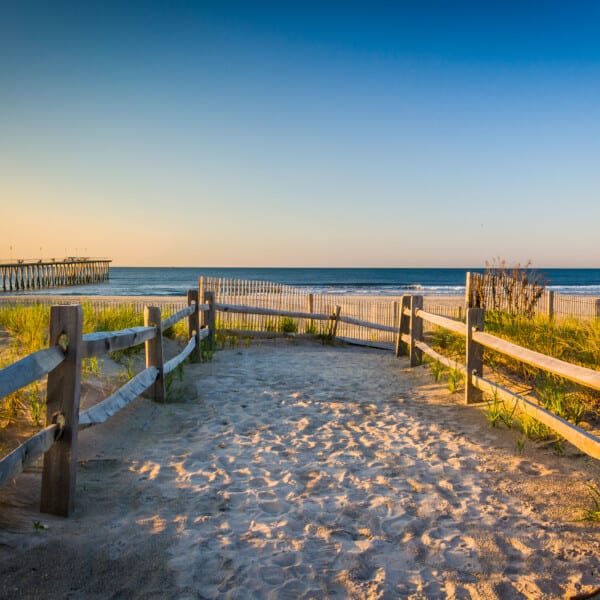 A serene beach pathway leading to a tranquil ocean view with a pier extending into the waters, under a vast, clear sky at dusk.