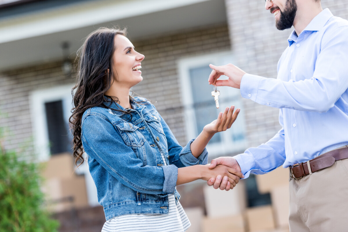 Happy woman receiving keys from a man in front of a house, symbolizing a new homeowner or real estate transaction.