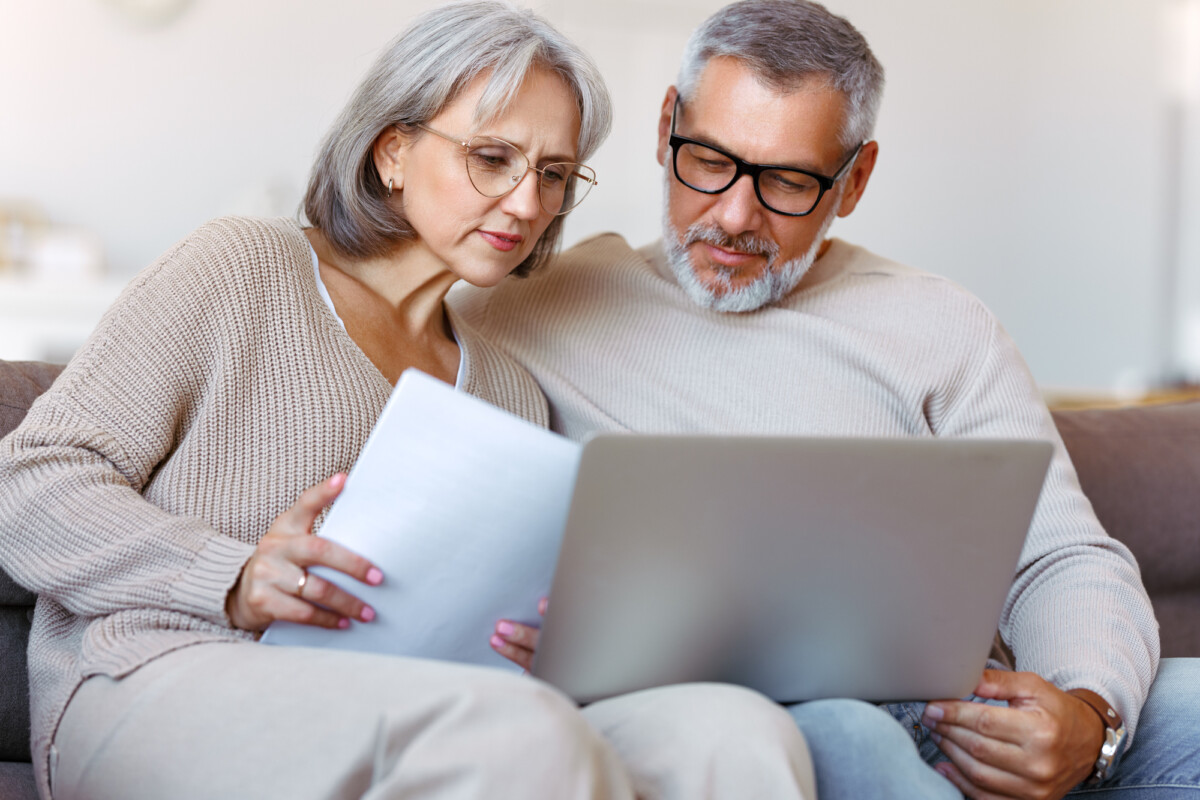 A mature couple comfortably sitting on a couch, reviewing documents together while using a laptop, suggesting a moment of collaboration and shared decision-making.