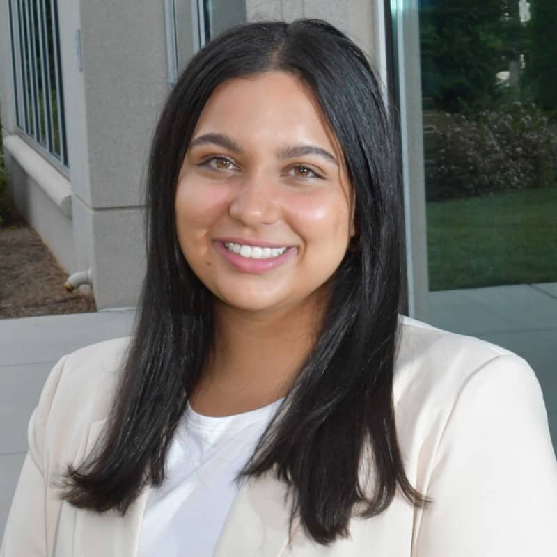 A professional portrait of Amani Sodhi. She is a smiling woman with long dark hair, dressed in a light beige blazer over a white top, posing outdoors with greenery and a building in the background.