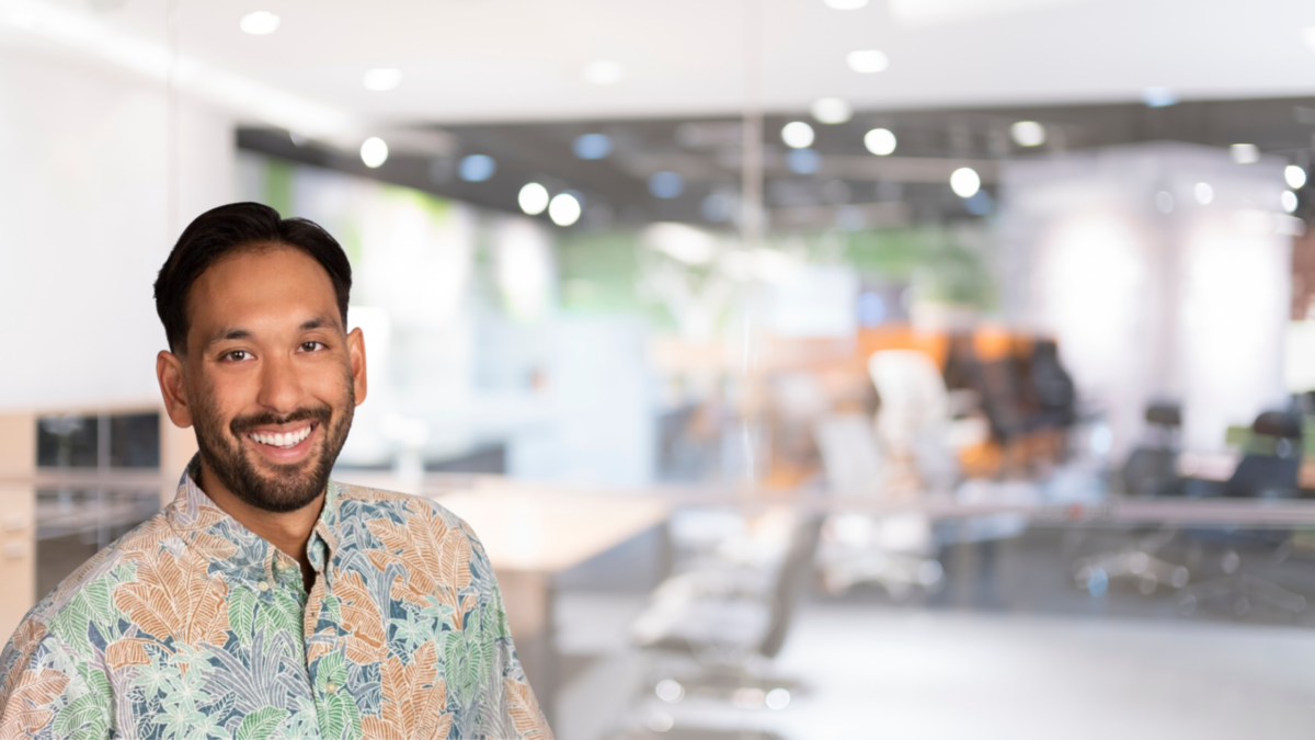 A smiling man with short dark hair and a beard is wearing a colorful floral shirt. He stands against a blurred background of a modern office with glass walls, office chairs, and an open workspace. The atmosphere appears bright and well-lit.