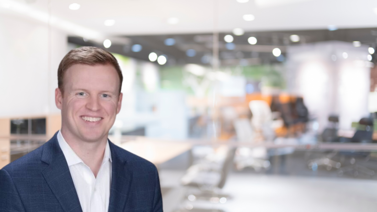 A man in a dark blue blazer smiles while standing in a bright, modern office setting. Out-of-focus office furniture and equipment are visible in the background, including chairs, desks, and computers.