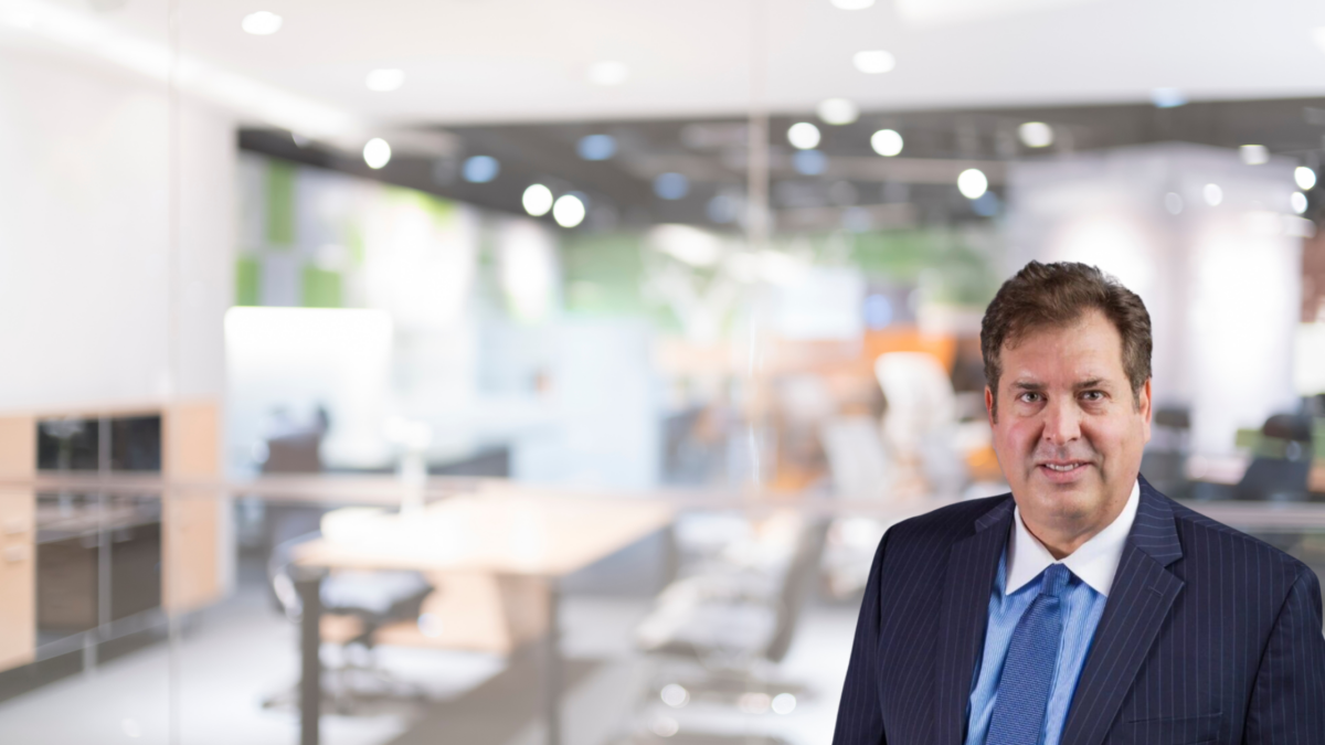 A middle-aged man in a blue suit and tie stands smiling in front of a modern, brightly lit office space with glass walls and blurred office furniture in the background.