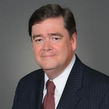 A middle-aged man, Bill Hortz, with short dark hair wearing a dark pinstripe suit, white dress shirt, and a maroon tie, posing against a plain gray backdrop. He has a slight smile and is looking directly at the camera.