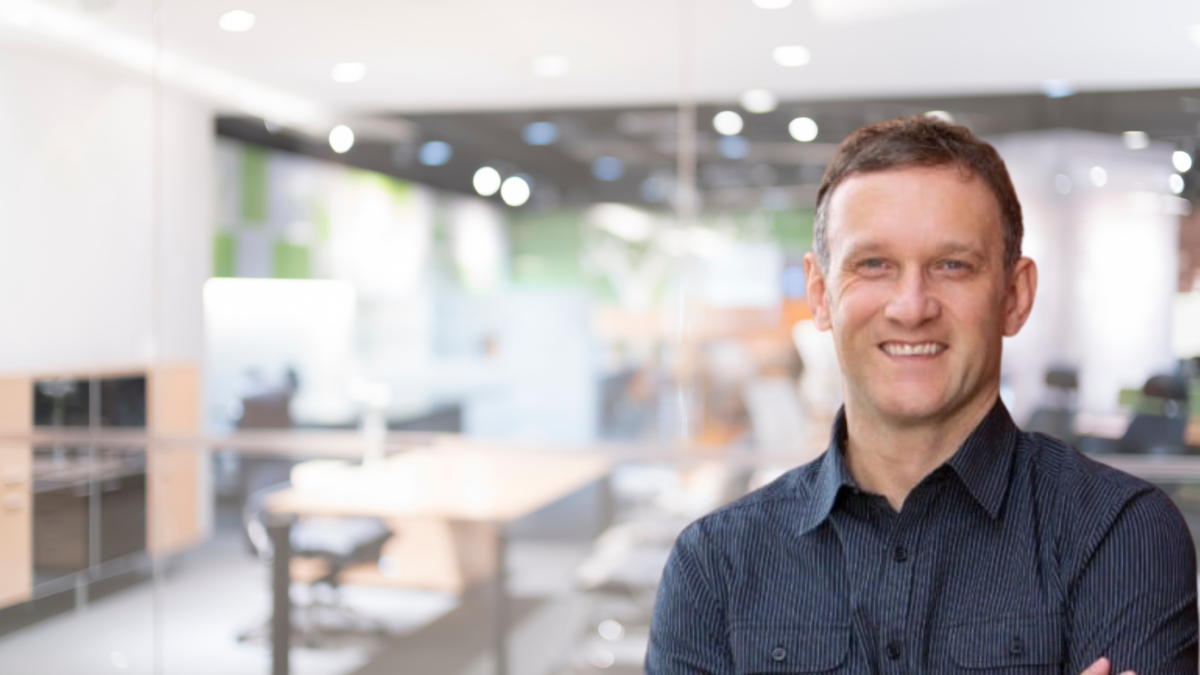 A man with short brown hair and a blue button-up shirt smiles with arms crossed in an office setting. The background features a modern office with desks, chairs, and bright lighting. The scene is slightly blurred, focusing on the man in the foreground.