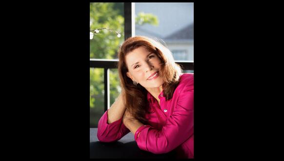 A woman with long brown hair smiles gently while resting her head on her hand. She is wearing a bright pink blouse and is seated in front of a window with leafy greenery visible outside. The sunlight softly illuminates her face.