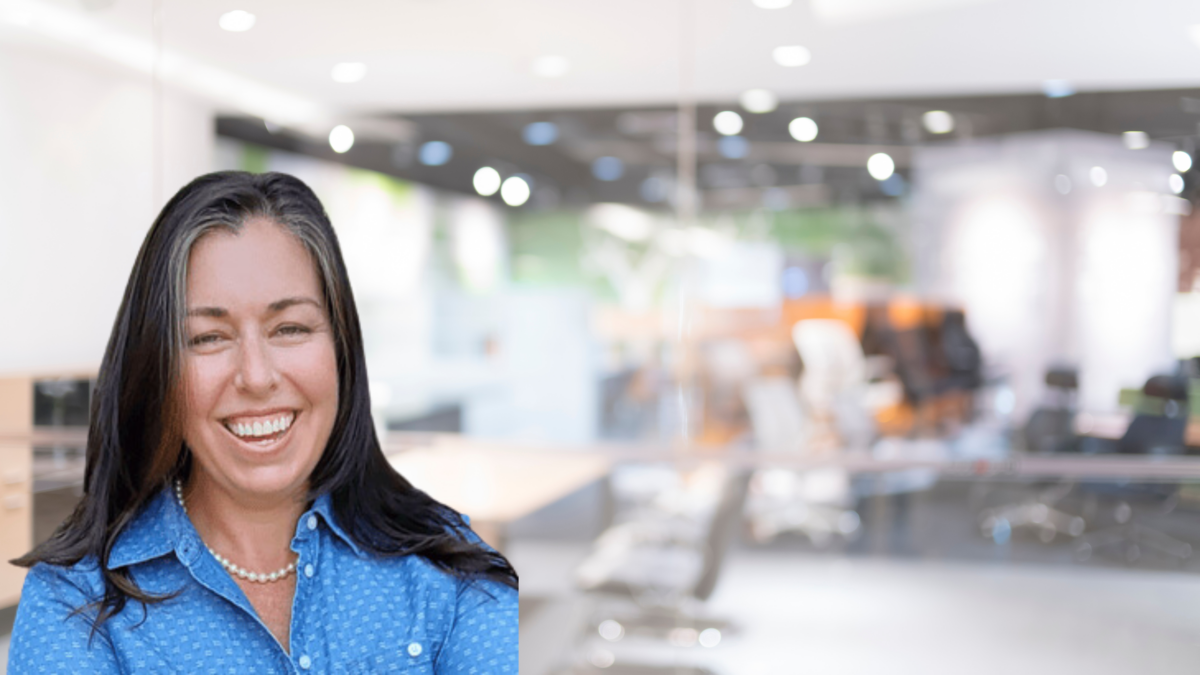 A woman with long dark hair and wearing a blue button-up shirt is smiling broadly. She is standing in a modern, brightly lit office space with glass walls, blurred office furniture and decor in the background, and numerous overhead lights.