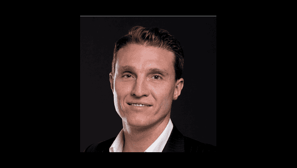 A man with short brown hair is smiling slightly. He is wearing a white shirt and a dark blazer. The background is black.