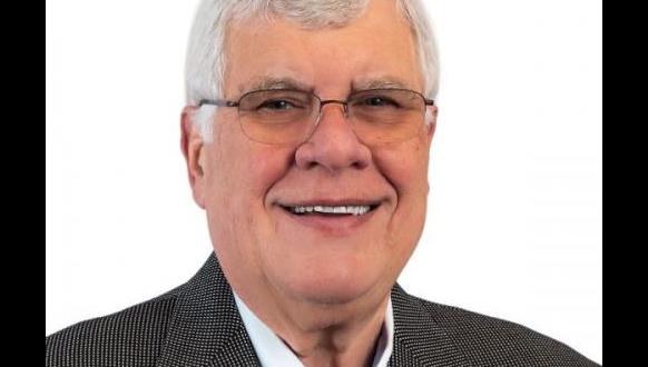 A person with short white hair and glasses smiles, wearing a gray checkered blazer and white shirt, against a plain white background.