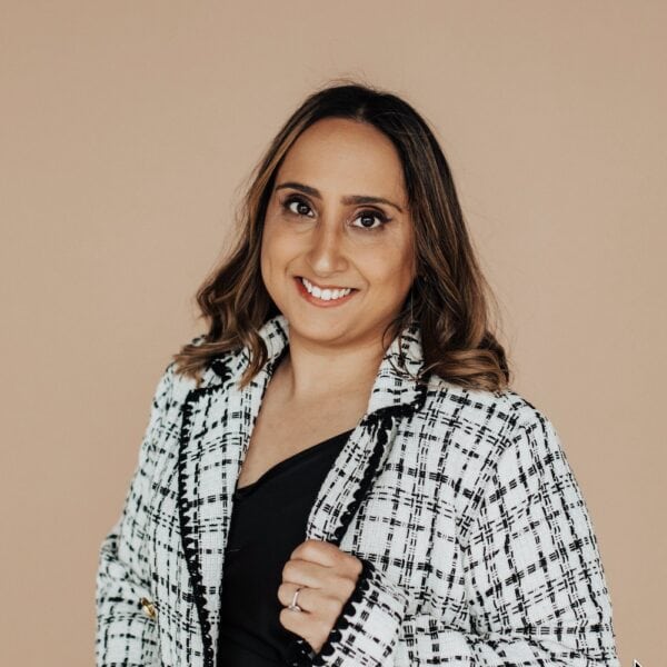 A person with shoulder-length brown hair is smiling, wearing a black top and a white and black checkered jacket. The background is a plain beige color.