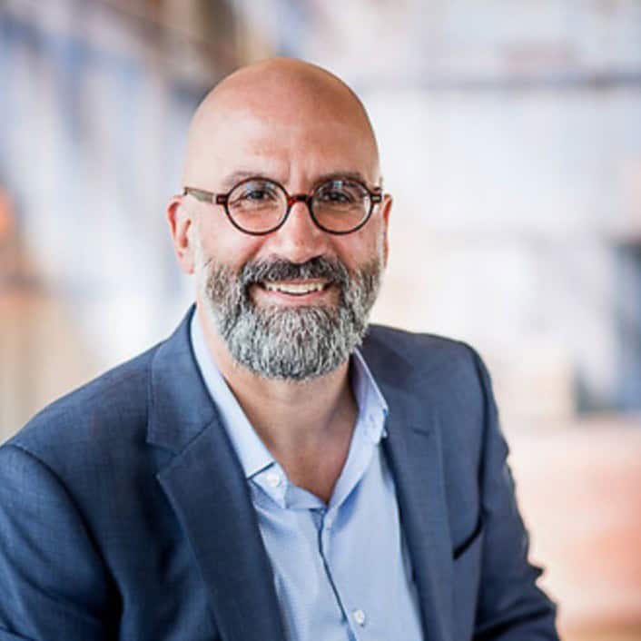 A smiling man with a bald head and a gray beard is wearing round glasses, a blue suit jacket, and a light blue shirt. The background is blurred, giving a soft focus effect.