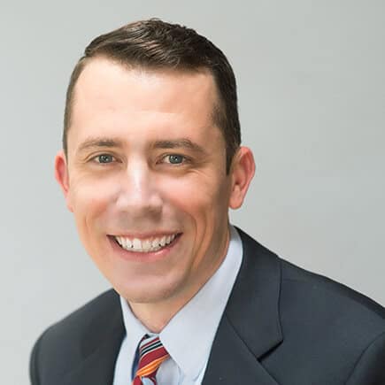 A man in a dark suit and a striped tie smiles at the camera against a plain background.