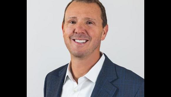 A person wearing a blue checkered blazer and a white shirt smiles at the camera against a plain background.