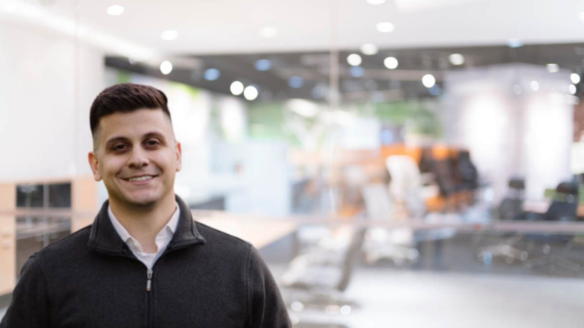 Smiling individual wearing a dark jacket stands in a modern office with a blurred background of desks and chairs under bright lighting.