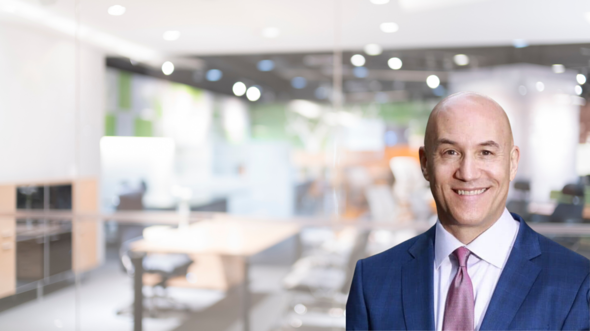 A bald man in a blue suit and pink tie smiles in an office setting. The background is blurred, showing a modern workspace with bright lighting and glass walls.