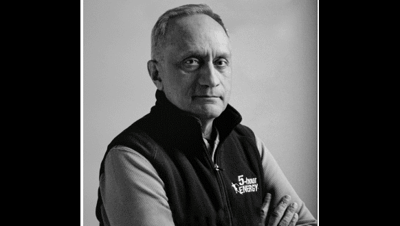 A man wearing a "5-hour Energy" vest stands against a plain background with his arms crossed. He looks directly at the camera, and his expression is neutral. The image is in black and white.