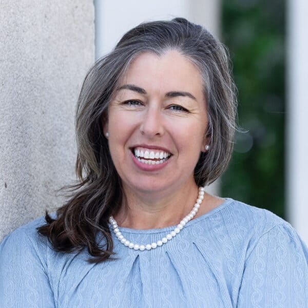 A woman with long brown and gray hair, wearing a light blue textured blouse and a pearl necklace, smiles while standing outdoors against a stone wall with greenery in the background.